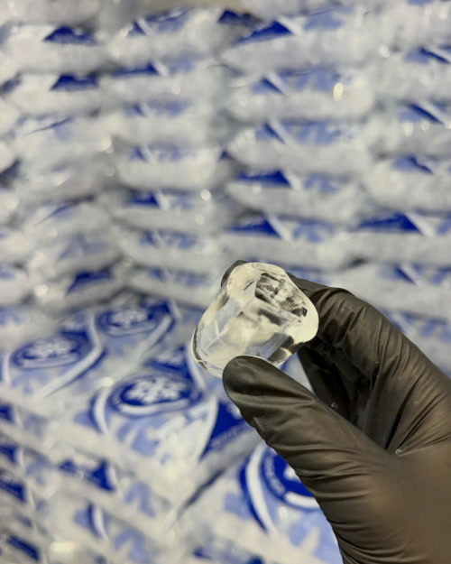 Mão com luva segurando um cubo de gelo cristalino da IcePoint, com as pilhas de pacotes ao fundo